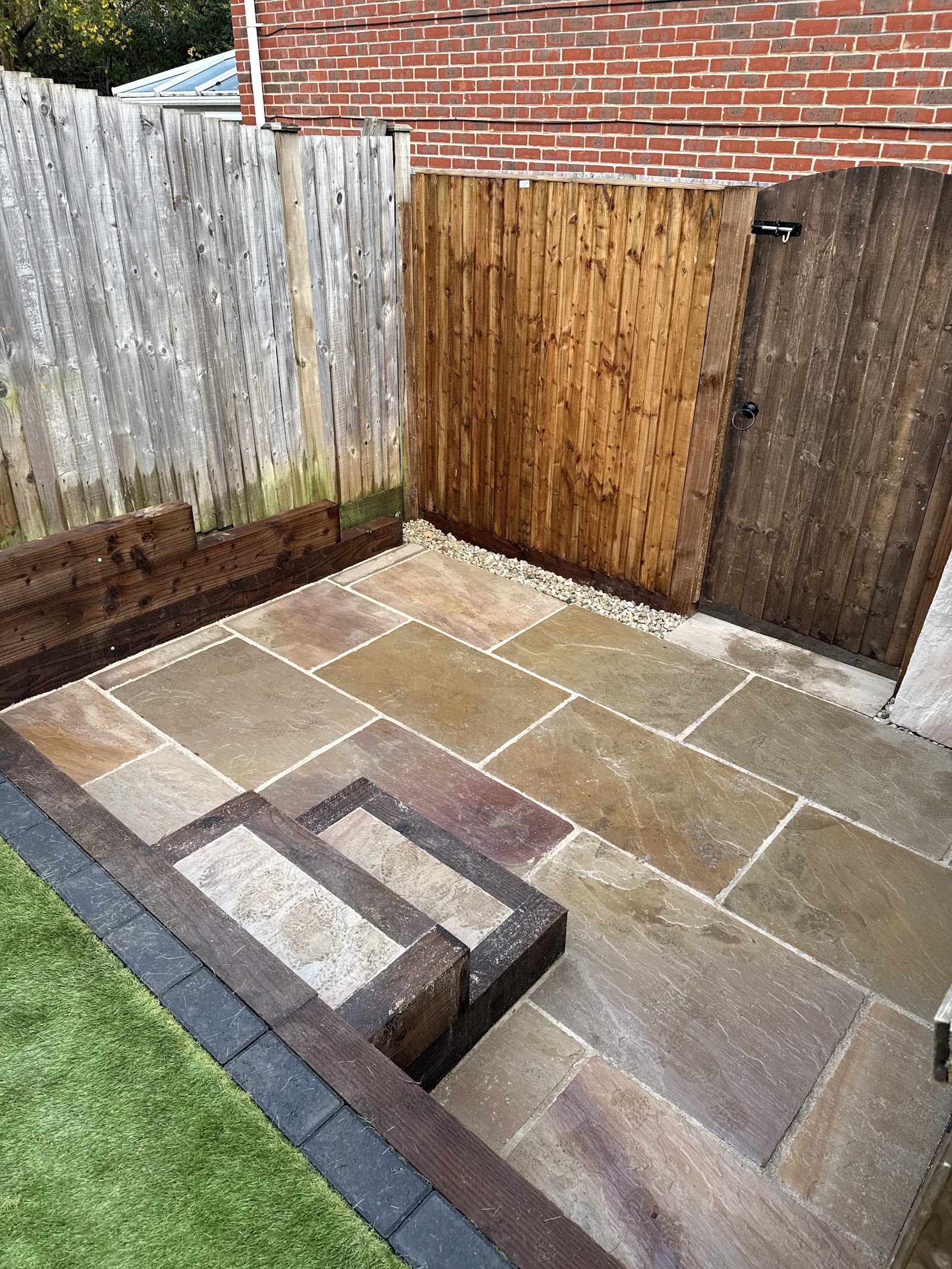 A riven Kandla-style Indian sandstone patio with a timber-sleeper edge and inset border, set against fresh larch-lap fencing and a new close-board gate.