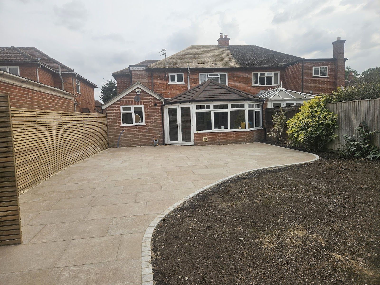 A large sand-toned porcelain terrace sweeping around the back of a semi-detached house, curved and bordered in setts, with beds left ready for planting.
