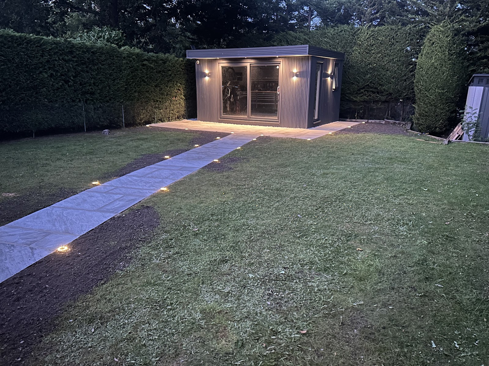 A natural-stone path lit low along its length running the depth of a lawn to a garden room at dusk — groundwork, paving and levels handled as one job.