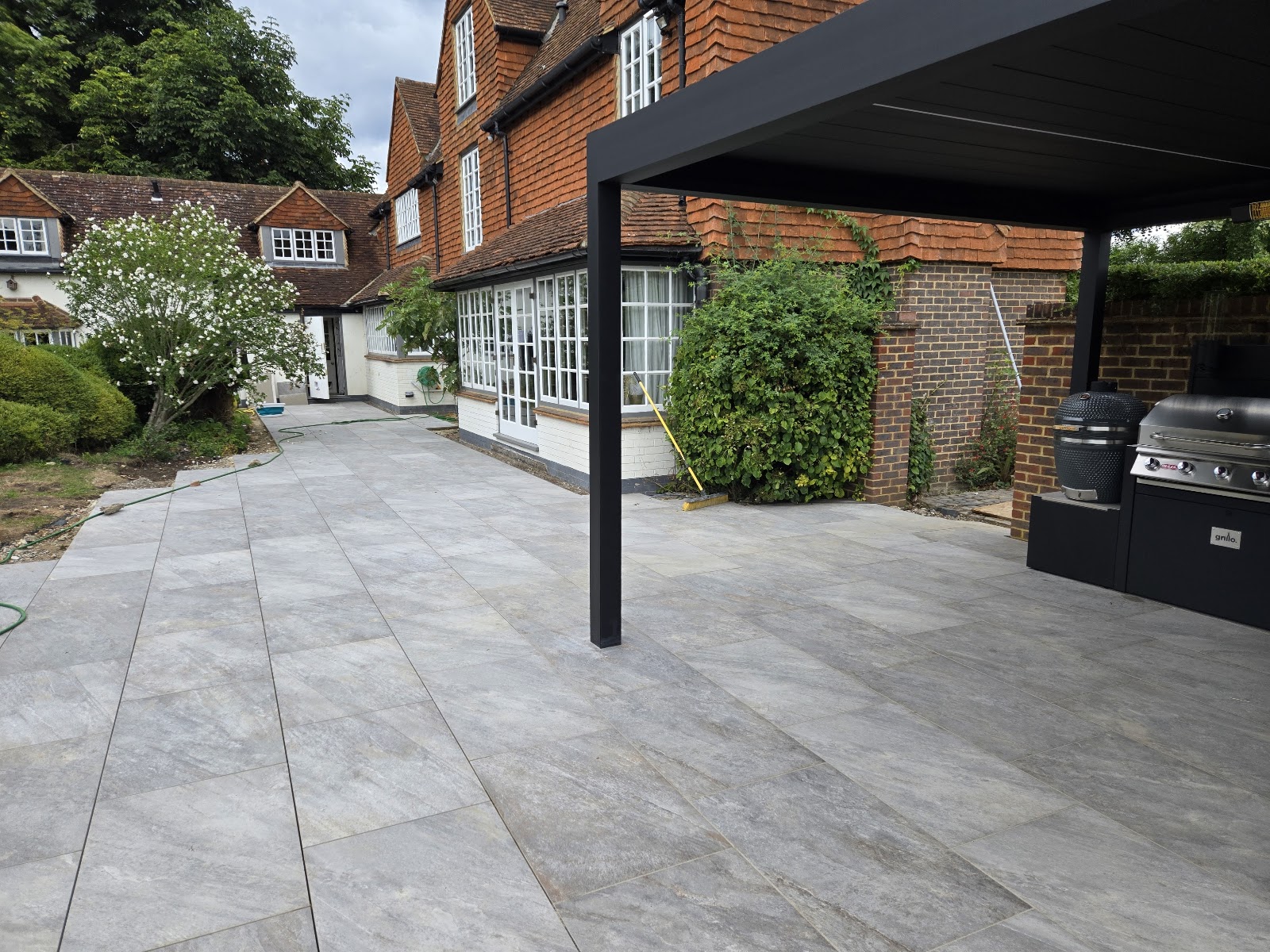 A wide grey porcelain courtyard behind a period tile-hung house, run out under a modern slatted pergola with an outdoor kitchen bay to one side.